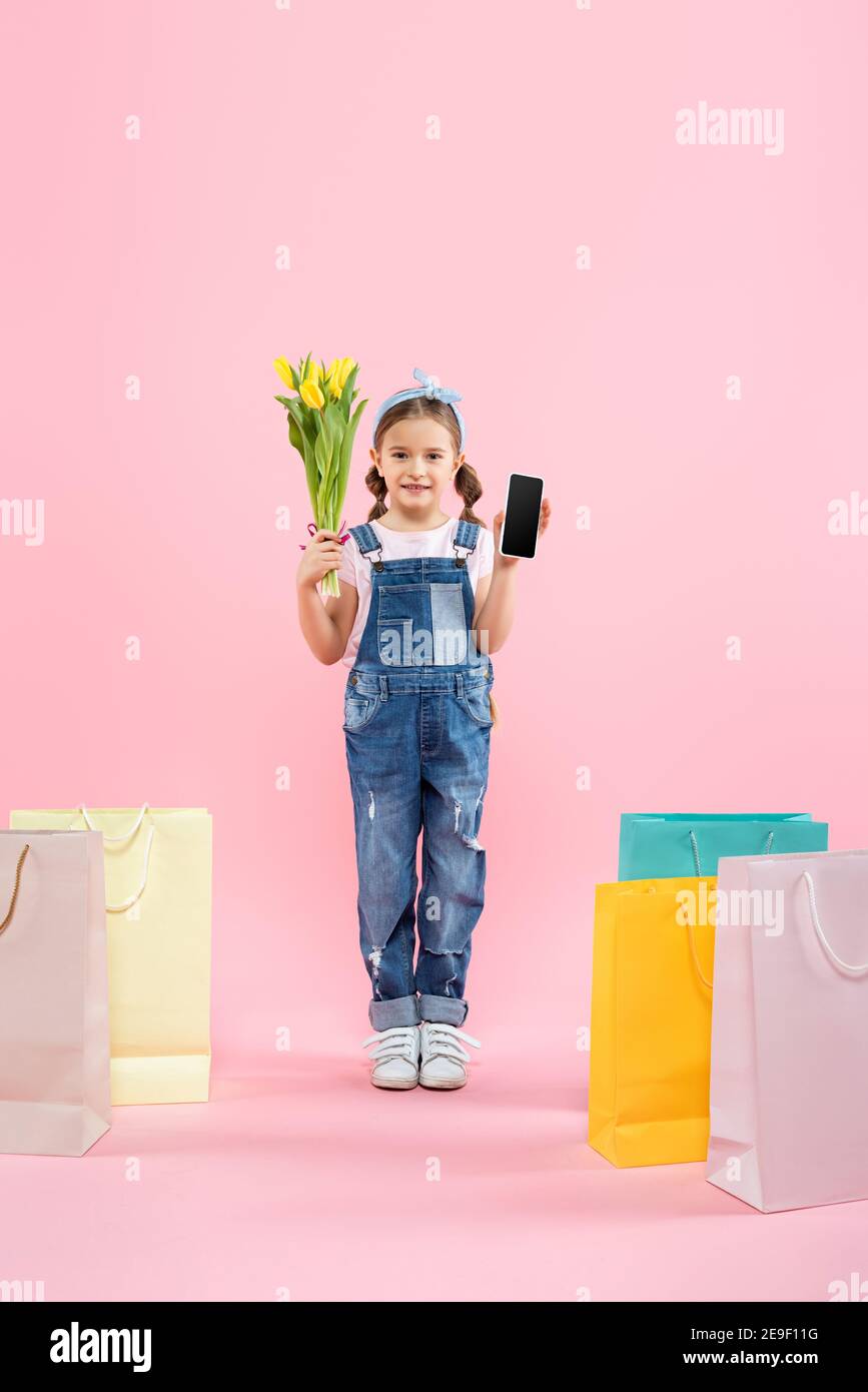 Volle Länge des Kindes hält Smartphone mit leerem Bildschirm und Tulpen in der Nähe von Einkaufstaschen auf rosa Stockfoto