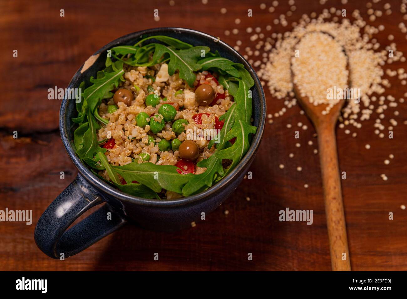 Nahaufnahme der veganen Quinoa-Salatschüssel auf Holztisch mit Rucola, Oliven, Erbsen, Tomaten. Rustikales gesundes hausgemachtes Gericht mit Holzlöffel Stockfoto