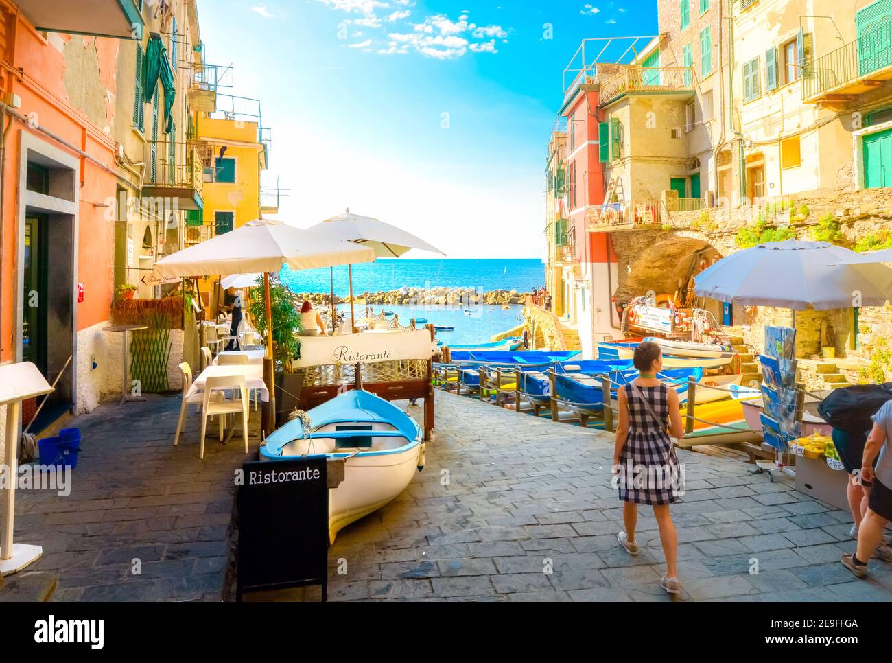Eine junge Frau im Karo-Kleid geht zur Bootsanlegestelle, vorbei an Straßencafés im bunten Dorf Cinque Terre in Riomaggiore, Italien. Stockfoto