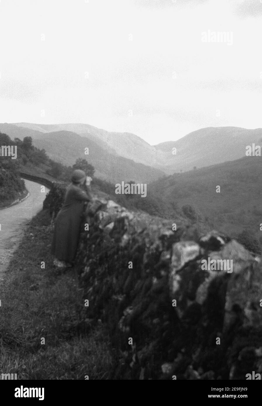 1930s, historisch, auf einem Grasrand, neben einer schmalen Landstraße auf einem Hügel, eine Dame mit Fernglas neben einer Wand, Blick über einen Valey, England, Großbritannien. Stockfoto