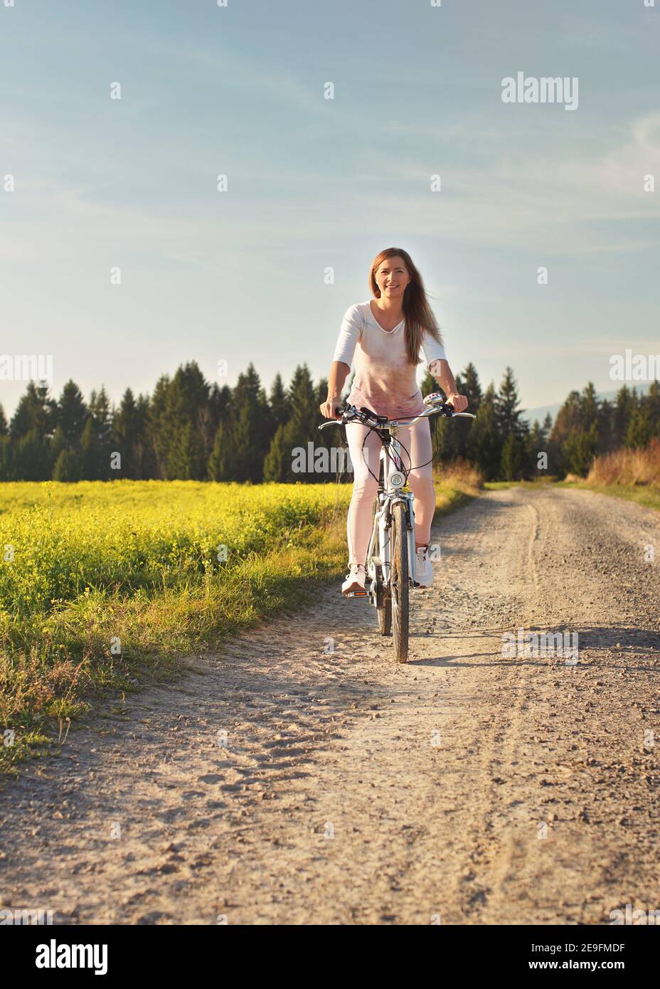 Junge Frau fährt Fahrrad auf staubigen Straße in Richtung Kamera, Nachmittag Sonne scheint auf Felder und Wald im Hintergrund Stockfoto