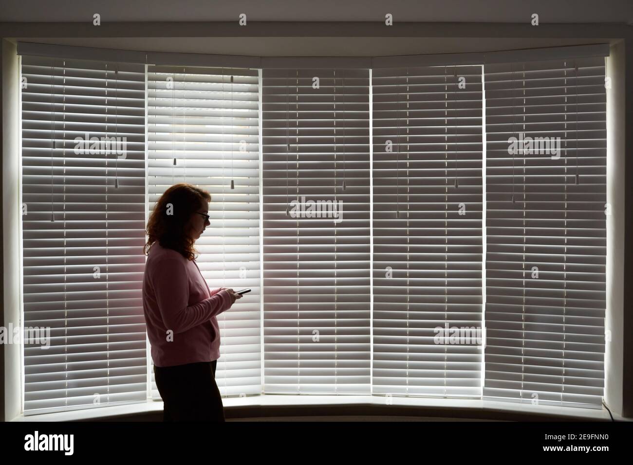 Foto einer jungen Frau in einer Shilouette, die während der Isolation des britischen Covid 19 vor einem großen Fenster stand. Frau hält ein Mobiltelefon. Stockfoto