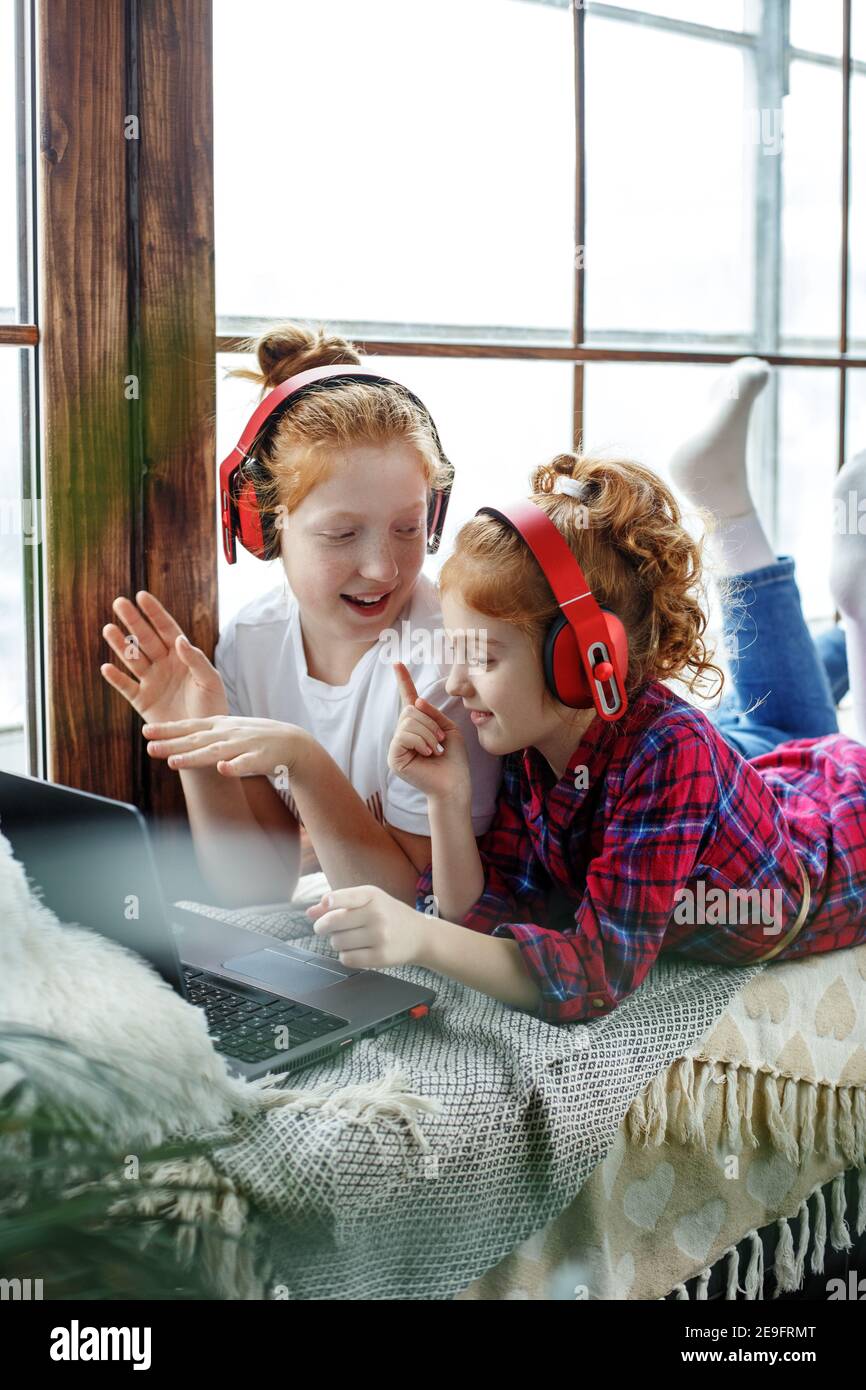 Zwei rothaarige Teenager-Mädchen in roten Kopfhörern hören fröhlich Musik, neben einem Laptop, sitzen auf dem Fenster Stockfoto