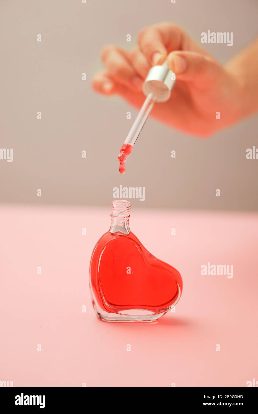 Herzförmige transparente Flasche auf einer rosa Oberfläche, eine Hand Tropfen einer roten Flüssigkeit mit einem Tropfer füllen die Flasche. Portrait-Modus Stockfoto