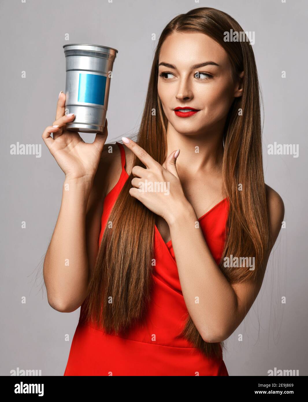 Junge Frau mit langen seidigen Haaren in roten Kleid zeigt Finger am Behälter mit Haarpflege Behandlung Kosmetik hält sie Stockfoto