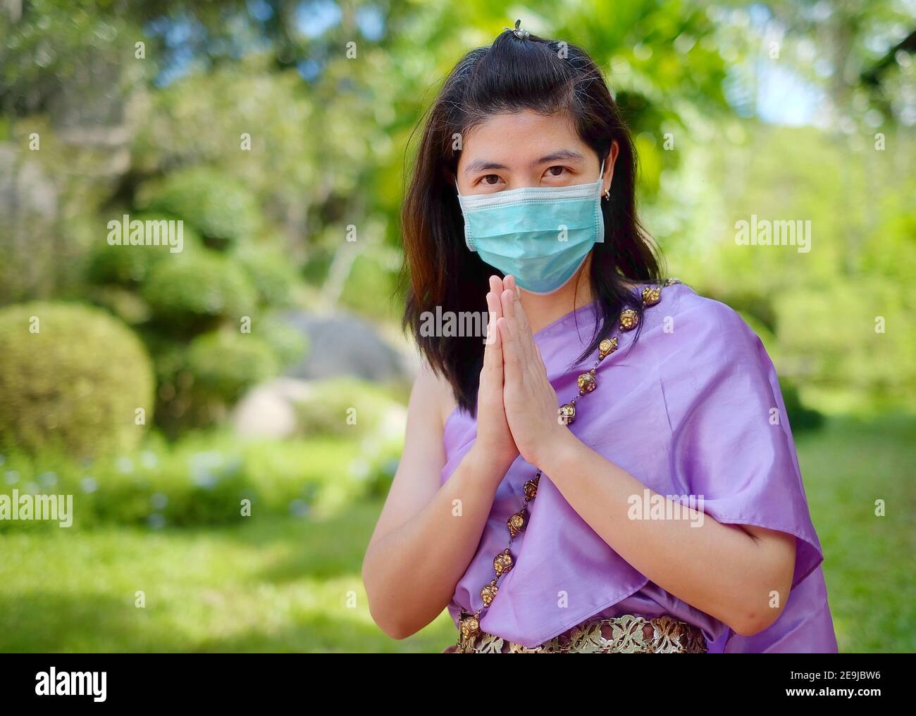 Eine Thailänderin in traditioneller Thai-Tracht, die eine OP-Maske trägt, zeigt einen thailändischen Grußruf "Wai", indem sie ihre Hände zusammenlegt und sich lehnt Stockfoto