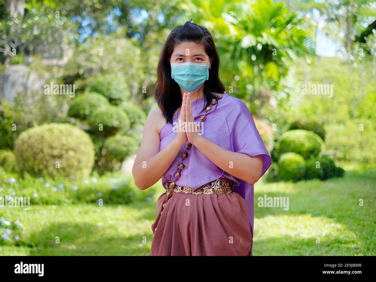 Eine Thailänderin in traditioneller Thai-Tracht, die eine OP-Maske trägt, zeigt einen thailändischen Grußruf "Wai", indem sie ihre Hände zusammenlegt und sich lehnt Stockfoto