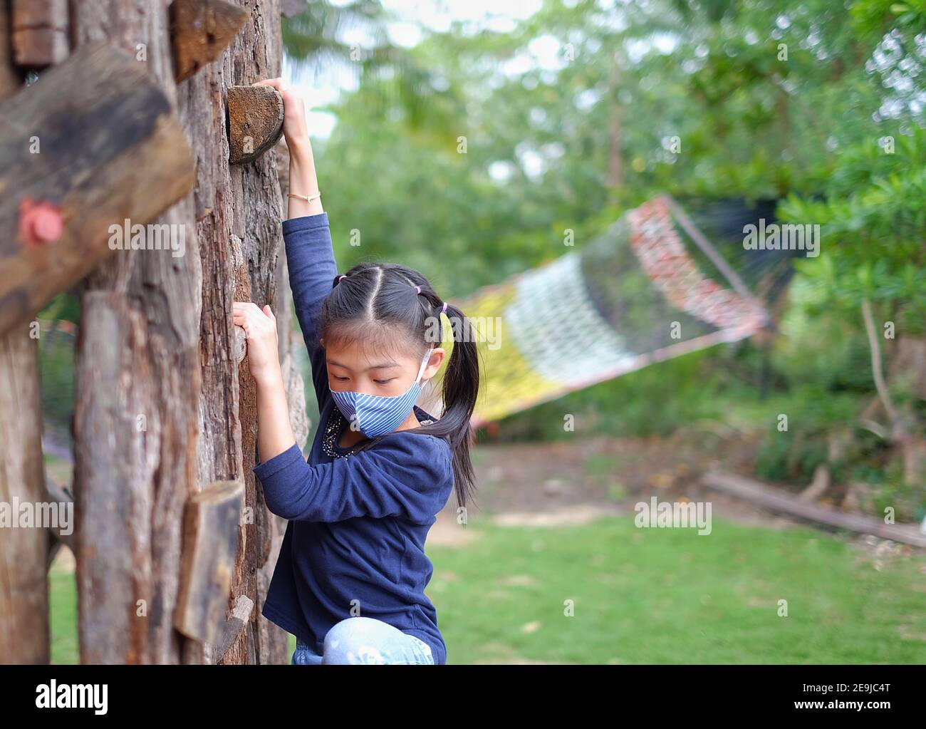 Ein nettes junges asiatisches Mädchen mit einer Gesichtsmaske klettert eine Wand eines Hindernisparcours, mutig, aber vorsichtig. Stockfoto