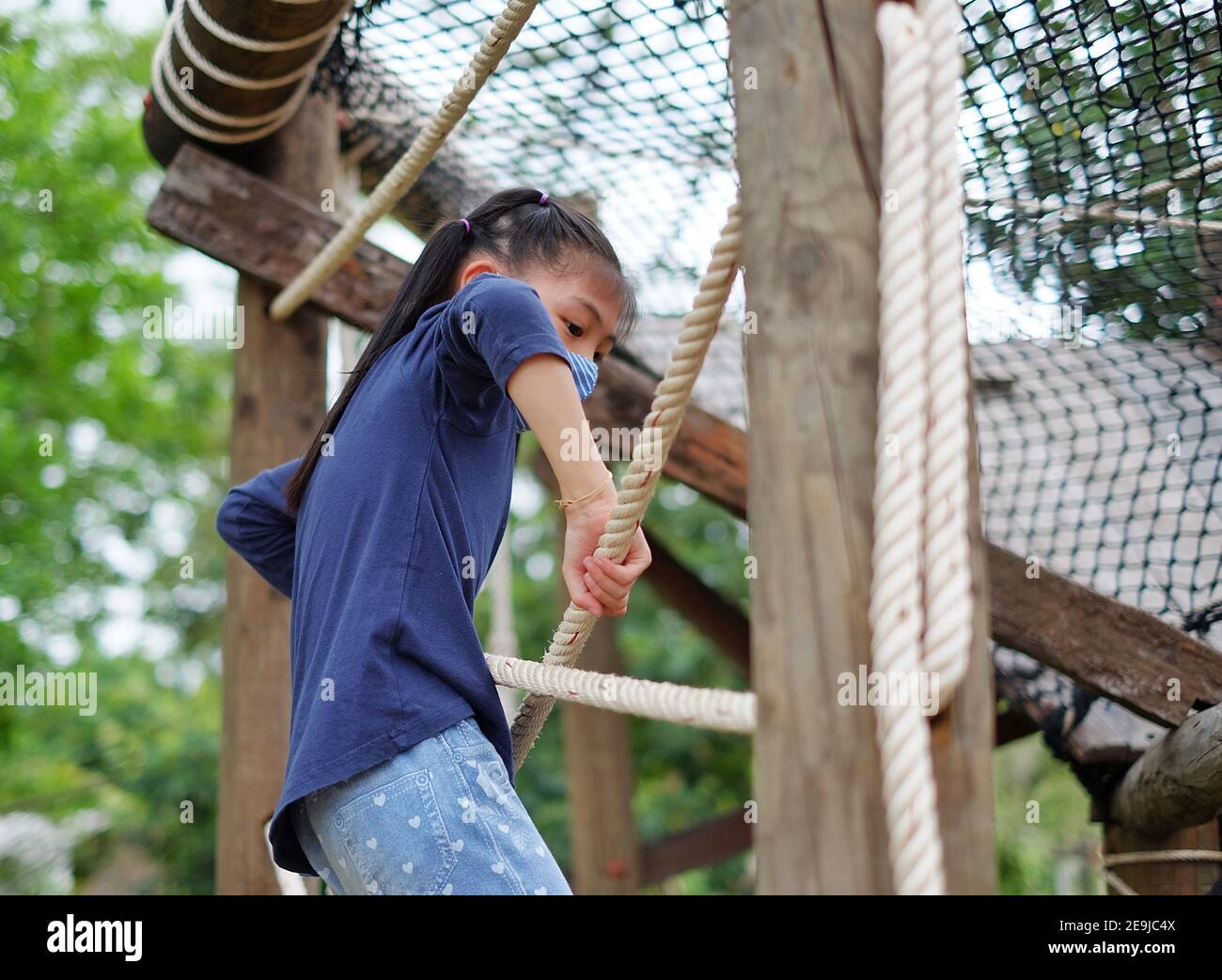 Ein nettes junges asiatisches Mädchen mit einer Gesichtsmaske klettert eine Wand eines Hindernisparcours, mutig, aber vorsichtig. Stockfoto