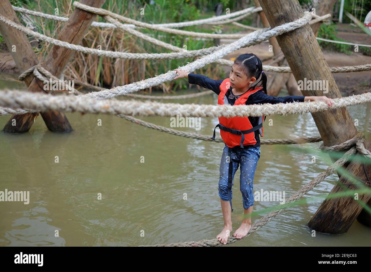 Ein süßes junges asiatisches Mädchen mit einer orangefarbenen Rettungsweste geht durch einen Hindernisparcours in einem Sommercamp mit einem engen Seil, das über dem Wasser hängt. Stockfoto
