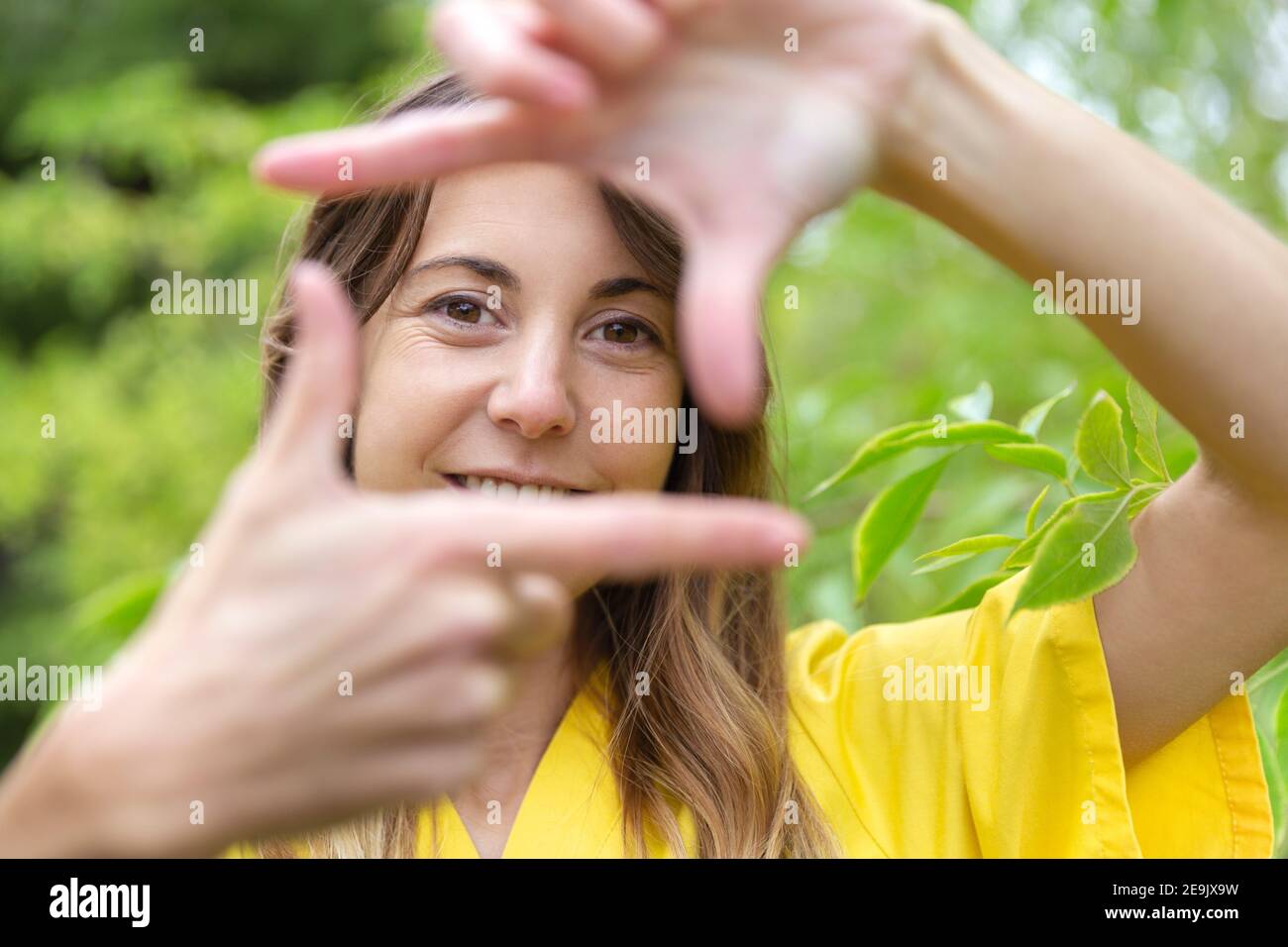 Nahaufnahme einer lächelnden jungen Frau, die ihr Gesicht mit ihren Händen als Selbstporträt umrahmt. Sie ist in der Natur. Leerzeichen für Text. Stockfoto