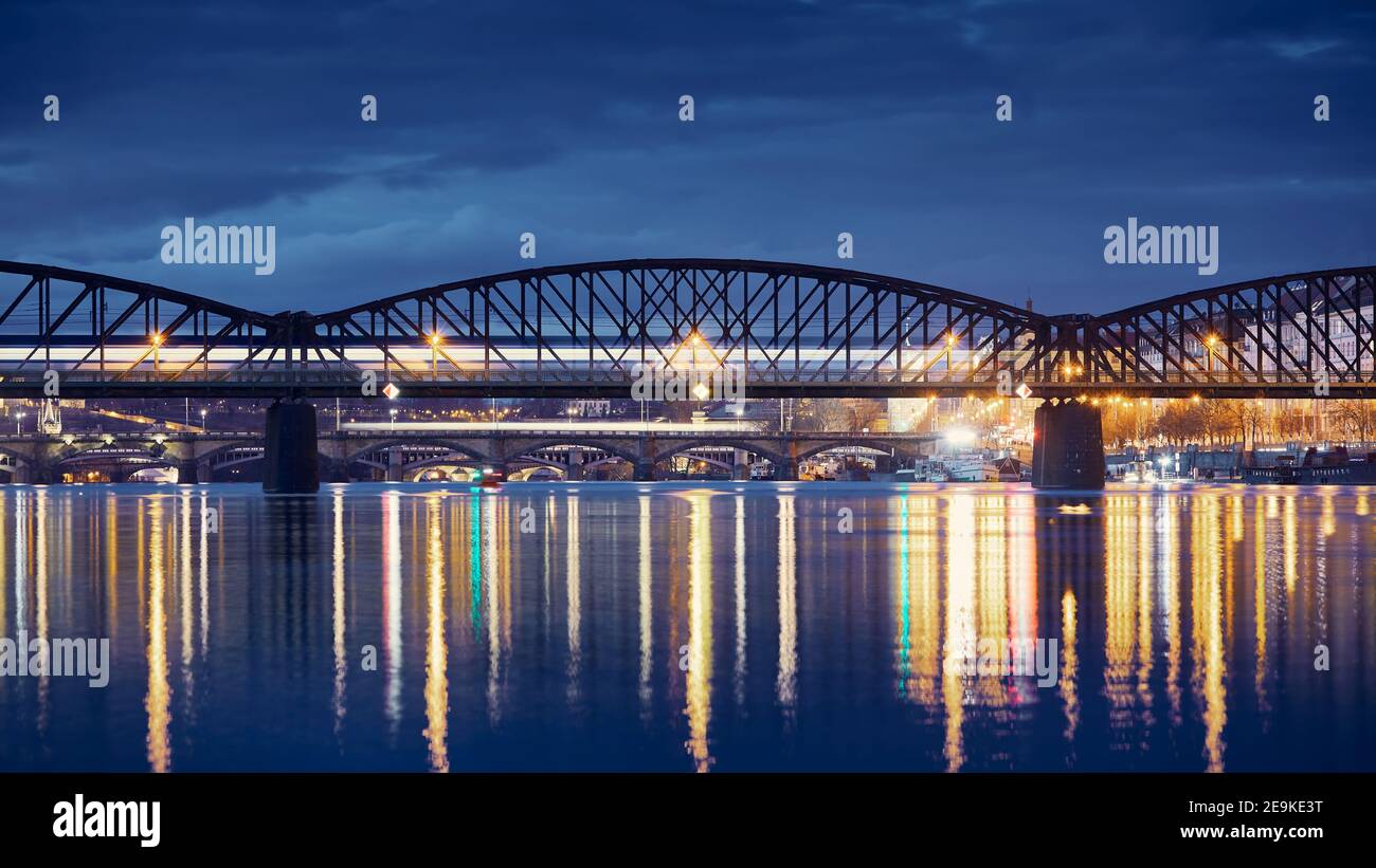 Personenzug, der über die Eisenbahnbrücke gegen die Stadt fährt. Stadtbild von Prag bei Nacht, Tschechische Republik Stockfoto
