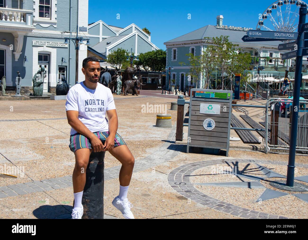 Waterfront - Kapstadt, Südafrika - 03-02-2021 stilvoller Mann, der auf einer Steinsäule an der Waterfront sitzt. Trendige Gebäude und Märkte im Hintergrund. Stockfoto