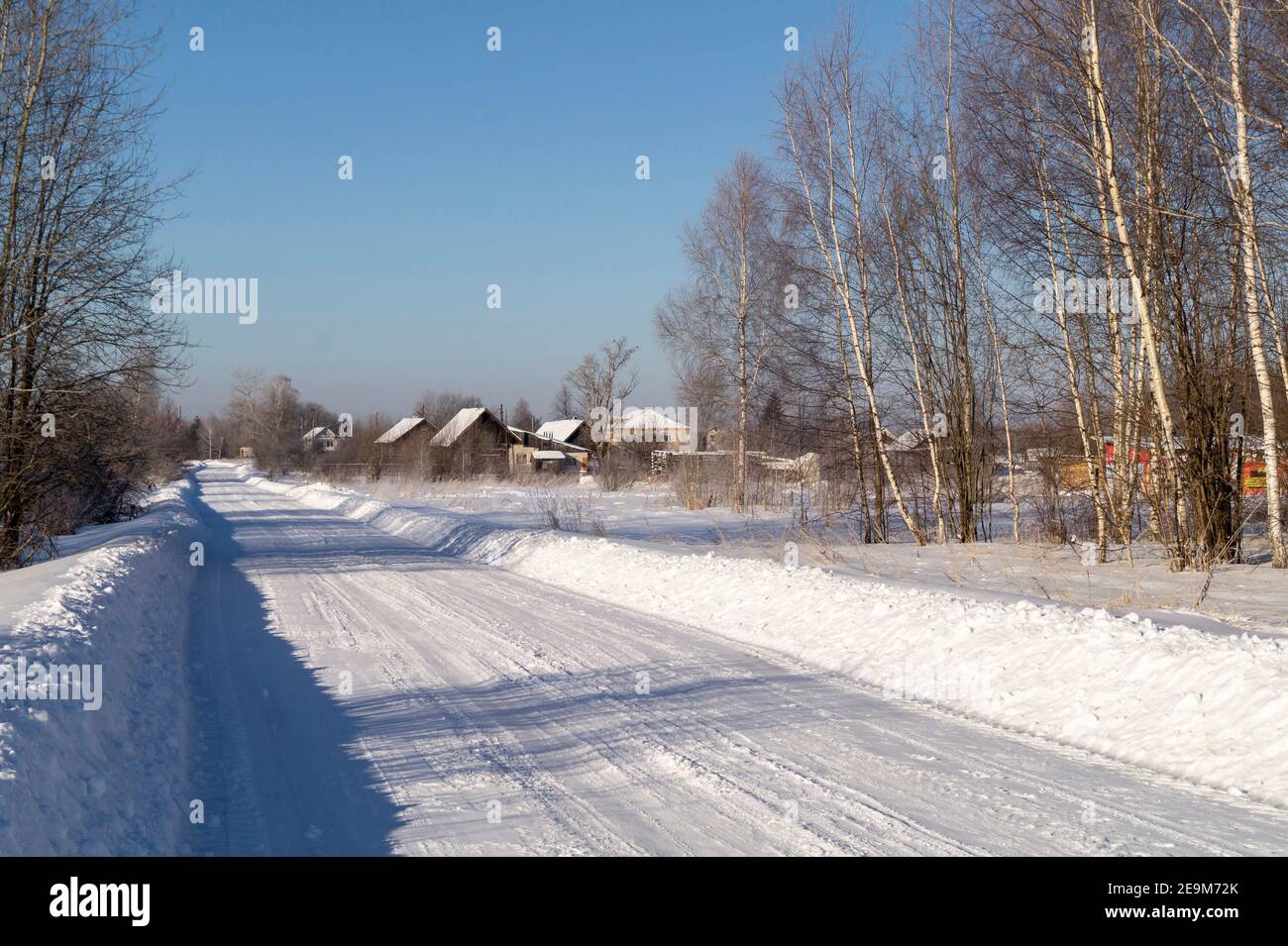 Die Winterstraße durch Feld im Dorf auf dem Hintergrund blauer Himmel. Schöne Landschaft in ländlichem Gelände bei Sonnentag Stockfoto