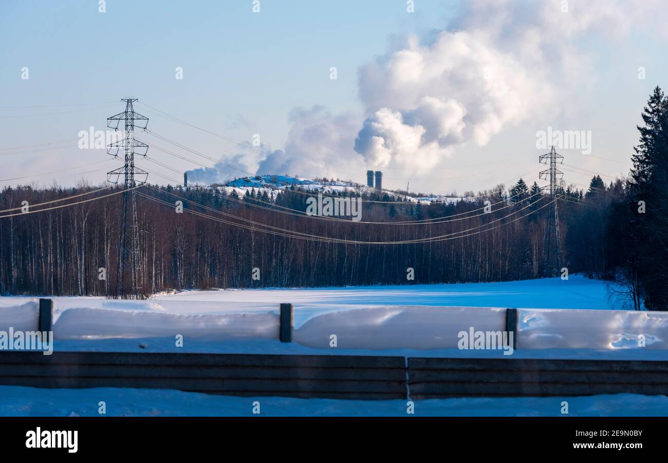 Helsinki / Finnland - 5. FEBRUAR 2021: Eine Stromleitung mit hohen Pylonen, die auf einem verschneiten Feld stehen. Zwei Kamine im Hintergrund. Stockfoto