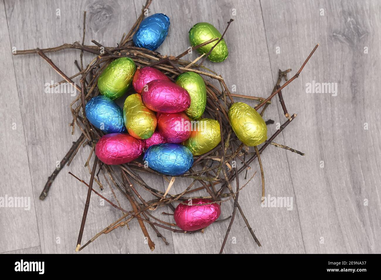 Folienumwickelte mehrfarbige ostereier in rosa, grün, blau und gelb in einem natürlichen Nest aus Stäben und Zweigen, vor einem grauen Holzhintergrund. Stockfoto