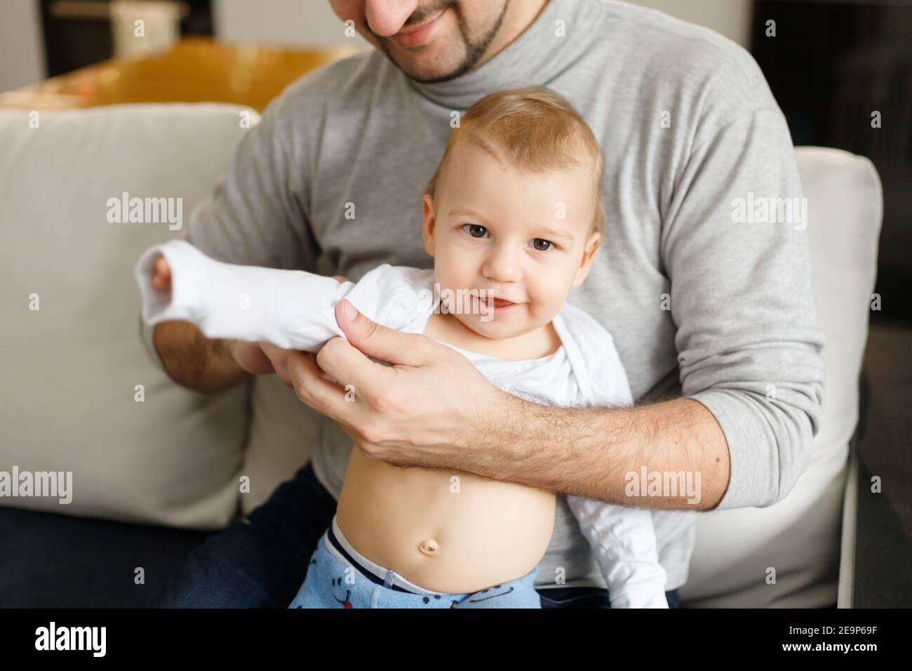 Vater kleidet Säugling im Wohnzimmer sitzen auf Couch zu Hause. Kleidung Baby Unterhemden und Slider auf Baby-Nahaufnahme. Home Care und Babypflege Konzept. Stockfoto