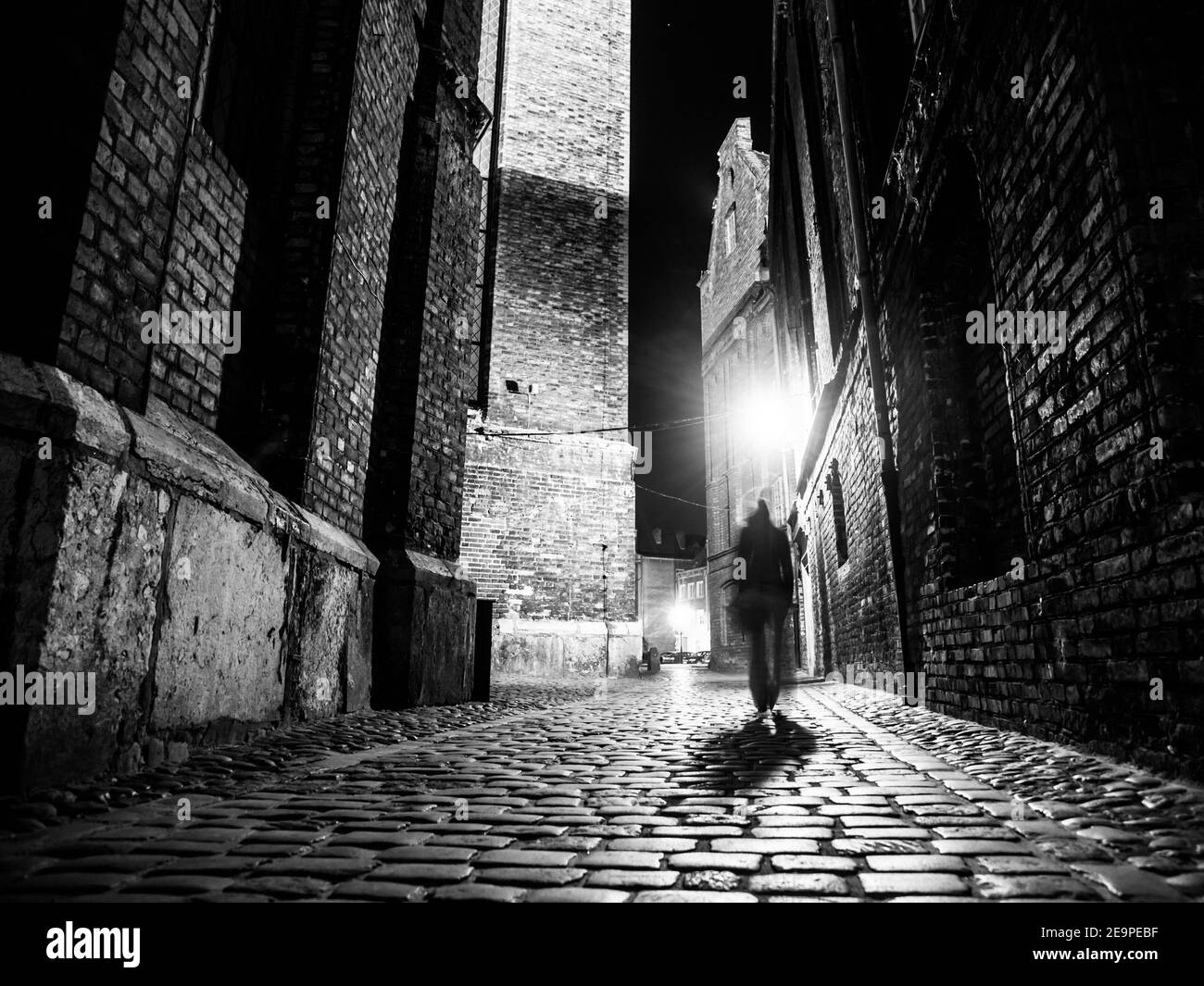 Beleuchtete gepflasterte Straße mit Lichtreflexen auf Kopfsteinpflaster in der alten historischen Stadt bei Nacht. Dunkle verschwommene Silhouette der Person erinnert an Jack the Ripper. Schwarzweiß-Bild. Stockfoto