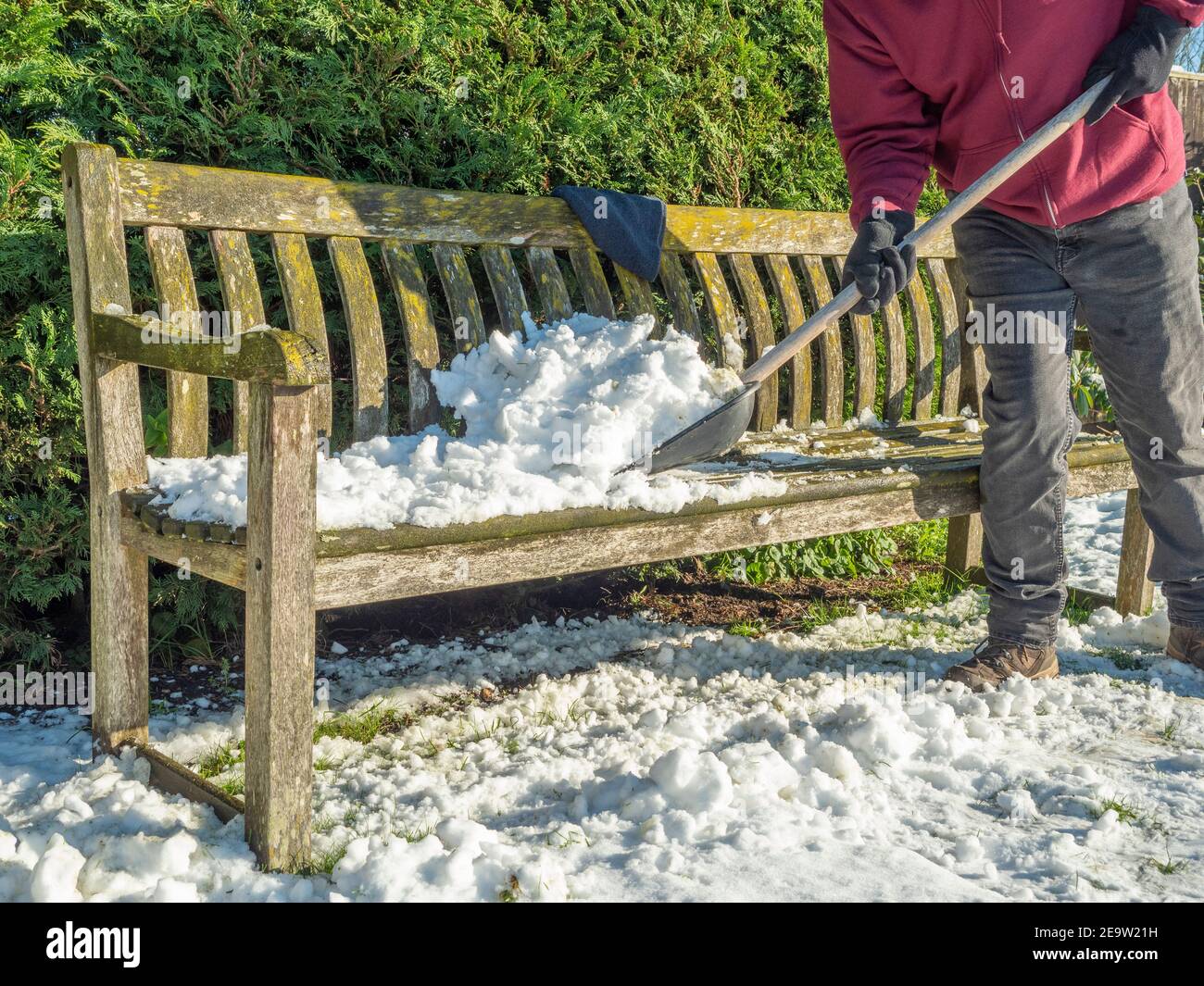 Nahaufnahme einer Person, die Stiefel und Handschuhe trägt, mit einer Schaufel, um eine Schicht Schnee vom Sitz eines Holzparks / Garten / Hof / Bank zu reinigen. Stockfoto