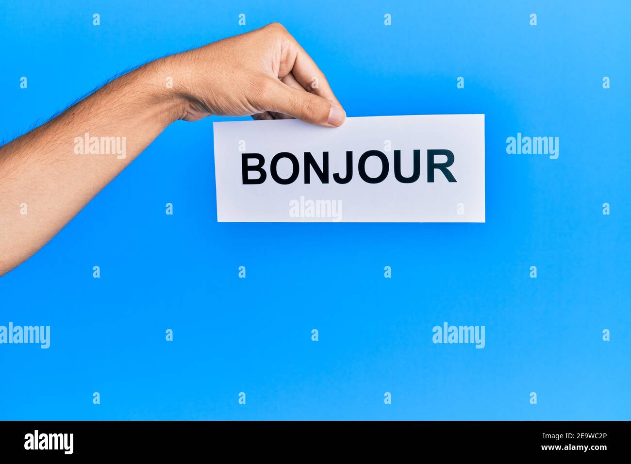 Hand von hispanischen Mann hält französisch Gruß bonjour Wort Papier über isolierten blauen Hintergrund. Stockfoto