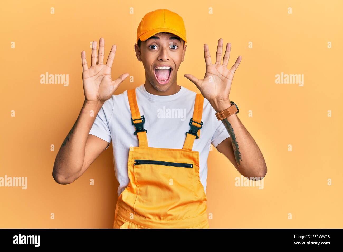 Junger, hübscher afroamerikanischer Mann, der eine Handyman-Uniform über Gelb trägt Hintergrund zeigt und zeigt nach oben mit den Fingern Zahl zehn während Lächelnde Konf Stockfoto