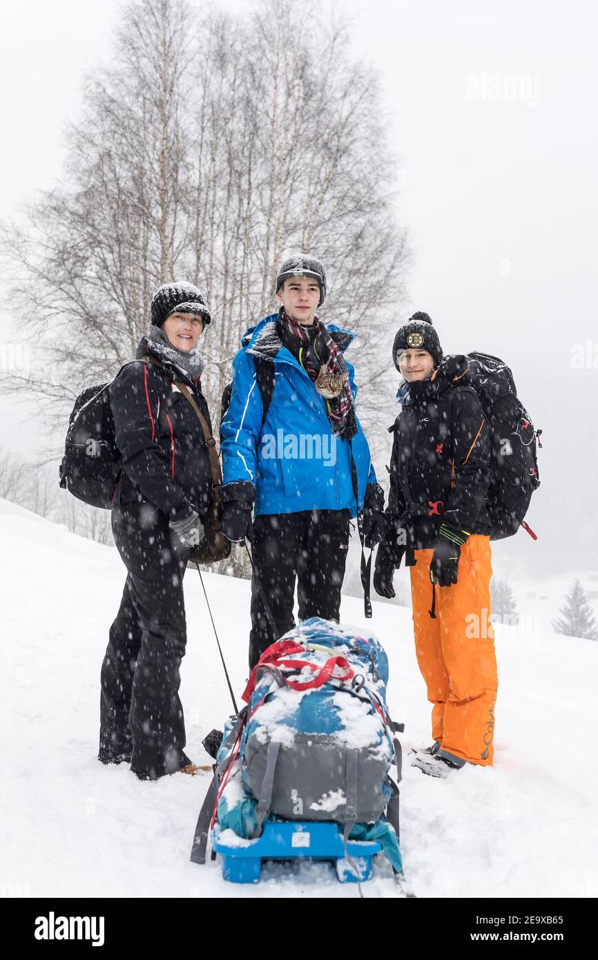 Drei Mitglieder Familie und ihre Katze, Mutter und zwei Söhne stehen mit ihren Rucksäcken und Gepäck in hügeliger Landschaft im Schneefall Stockfoto
