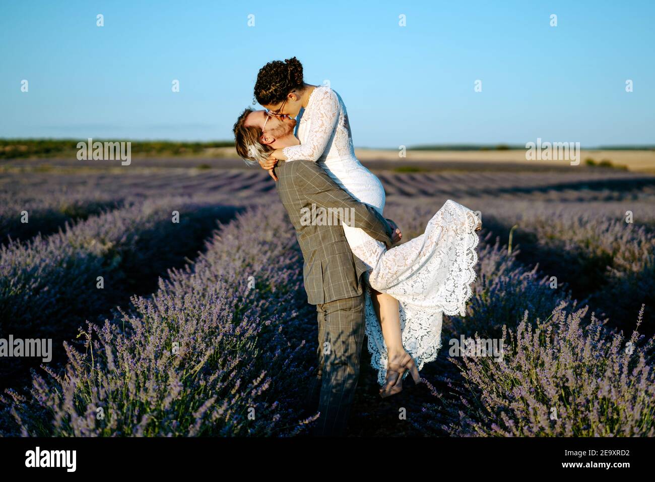 Seitenansicht des Bräutigams, der die Braut hebt, während er in Lavendel steht Feld auf dem Hintergrund des klaren blauen Himmels am Hochzeitstag Stockfoto