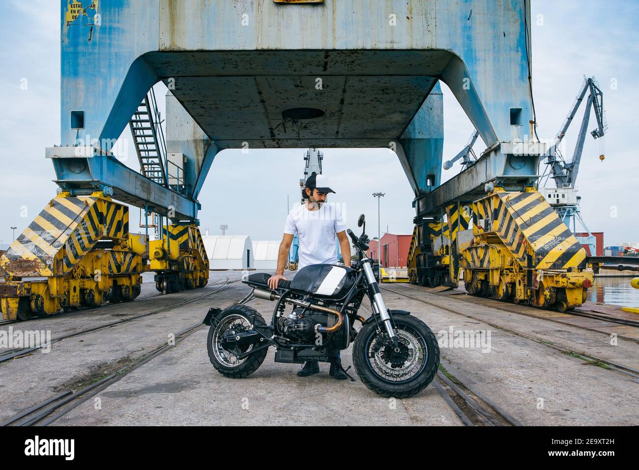 Ganzer Körper von schweren jungen ethnischen männlichen Biker in casual Kleidung und Kappe stehen in der Nähe von Motorrad in der Nähe von Industriekranen Am Meer Stockfoto