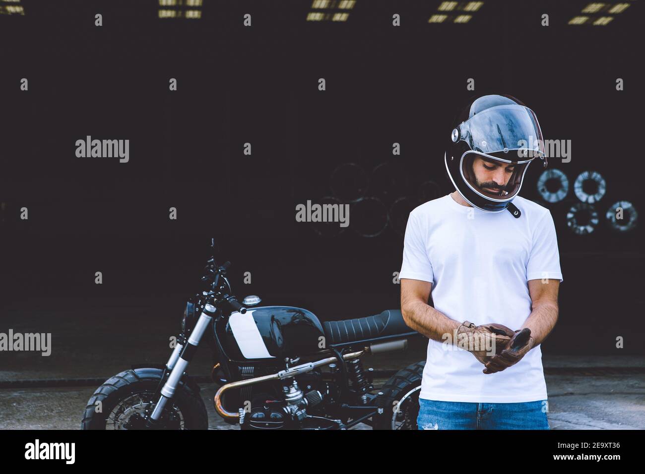 Ganzer Körper von schweren jungen ethnischen männlichen Biker in casual Kleidung und Kappe stehen in der Nähe von Motorrad in der Nähe von Industriekranen Am Meer Stockfoto
