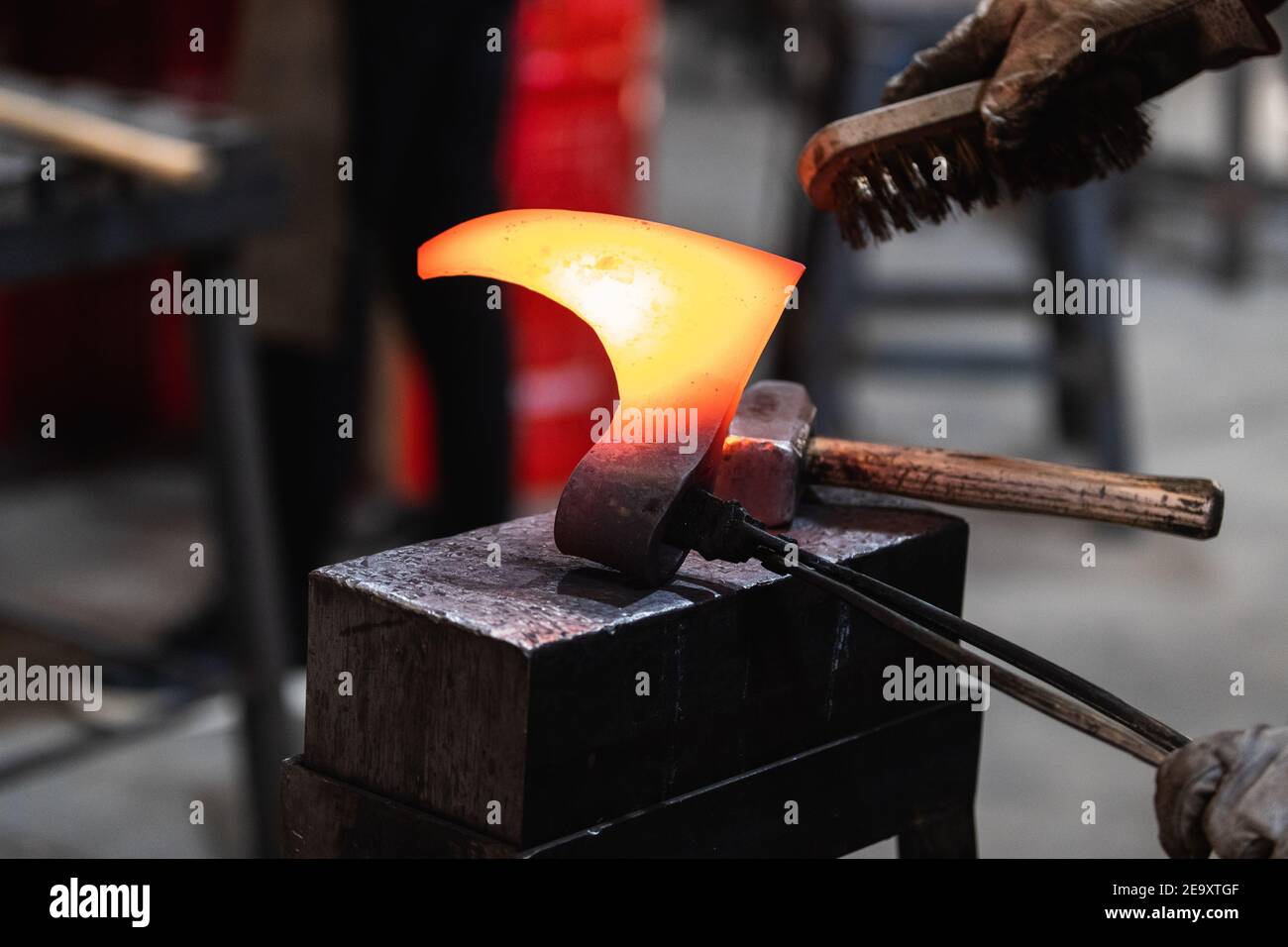 Crop gesichtslosen Schmied hält heißen Metall Detail der wikinger Axt Mit Zangen bei der Arbeit mit Drahtbürste in Schmiede Stockfoto