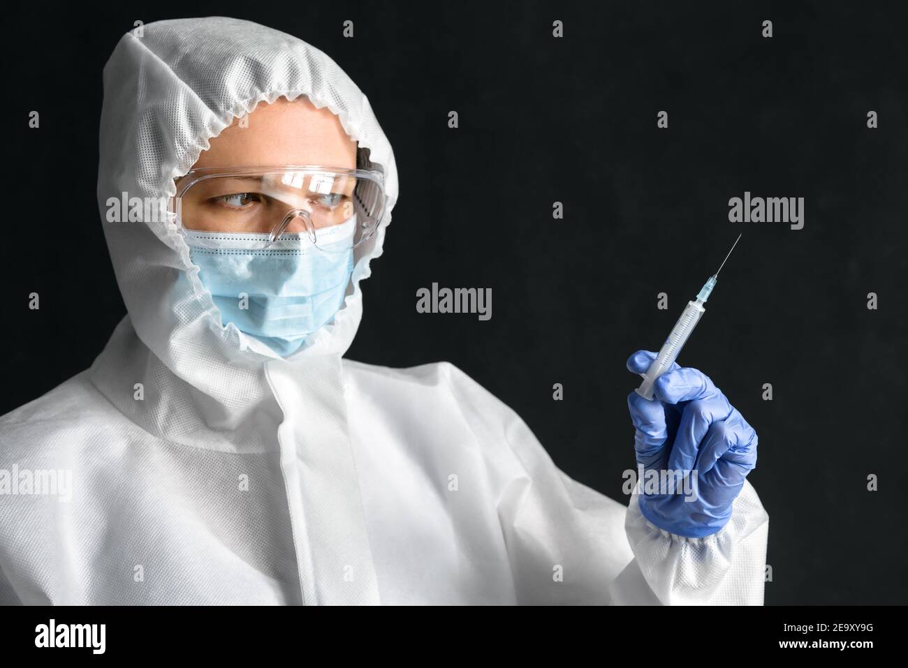 Krankenschwester in persönlicher Schutzausrüstung (PSA) hält Spritze für COVID-19-Impfstoffinjektion. Porträt des Arztes trägt Gesichtsmaske und Brille auf dunklen BA Stockfoto