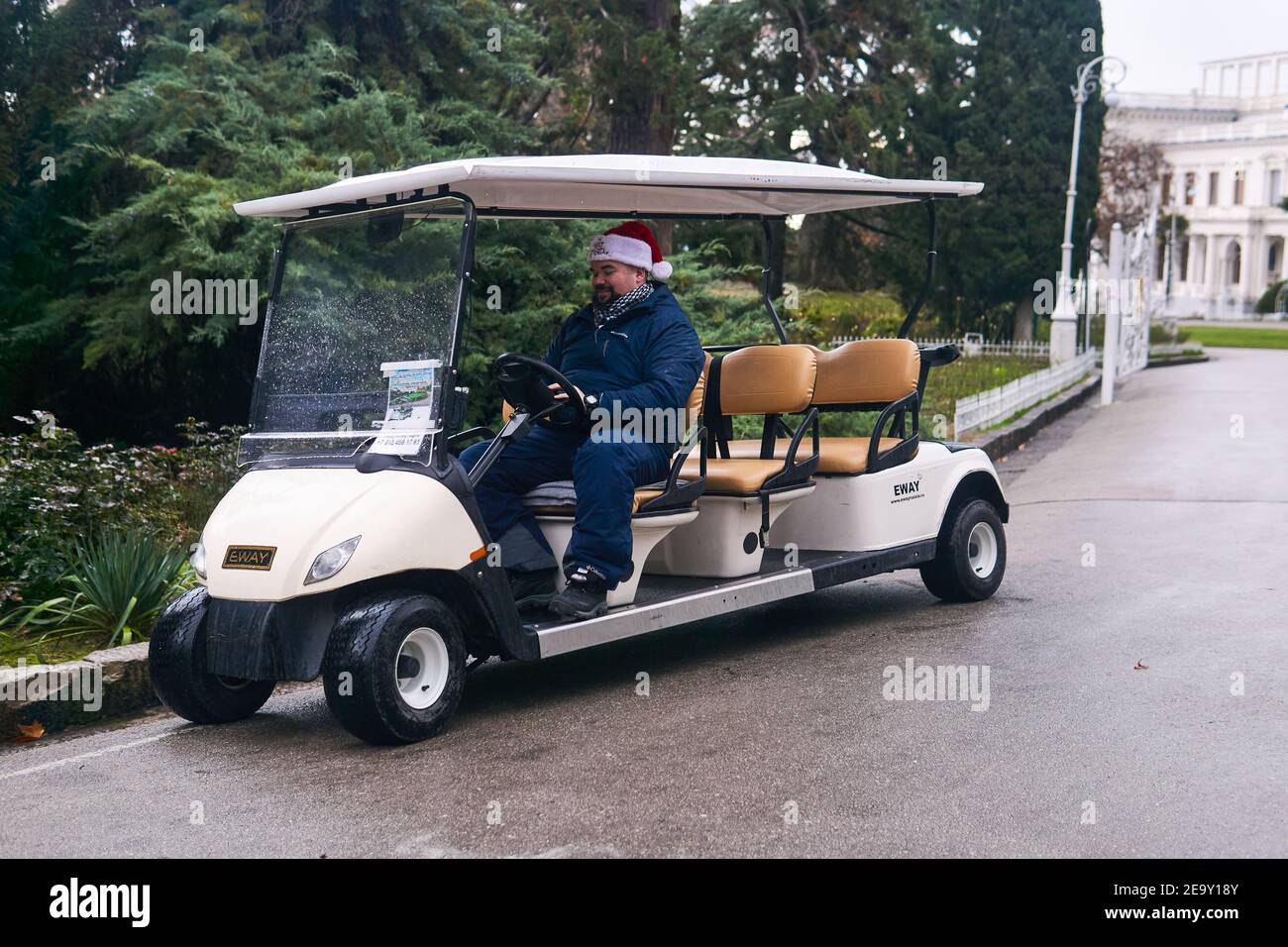 Jalta, Russland - 26. Januar 2021: Fahrer eines touristischen elektrischen Golfwagens in einem Weihnachtshut schaut in ein Smartphone und lächelt in seiner Freizeit in der Stockfoto