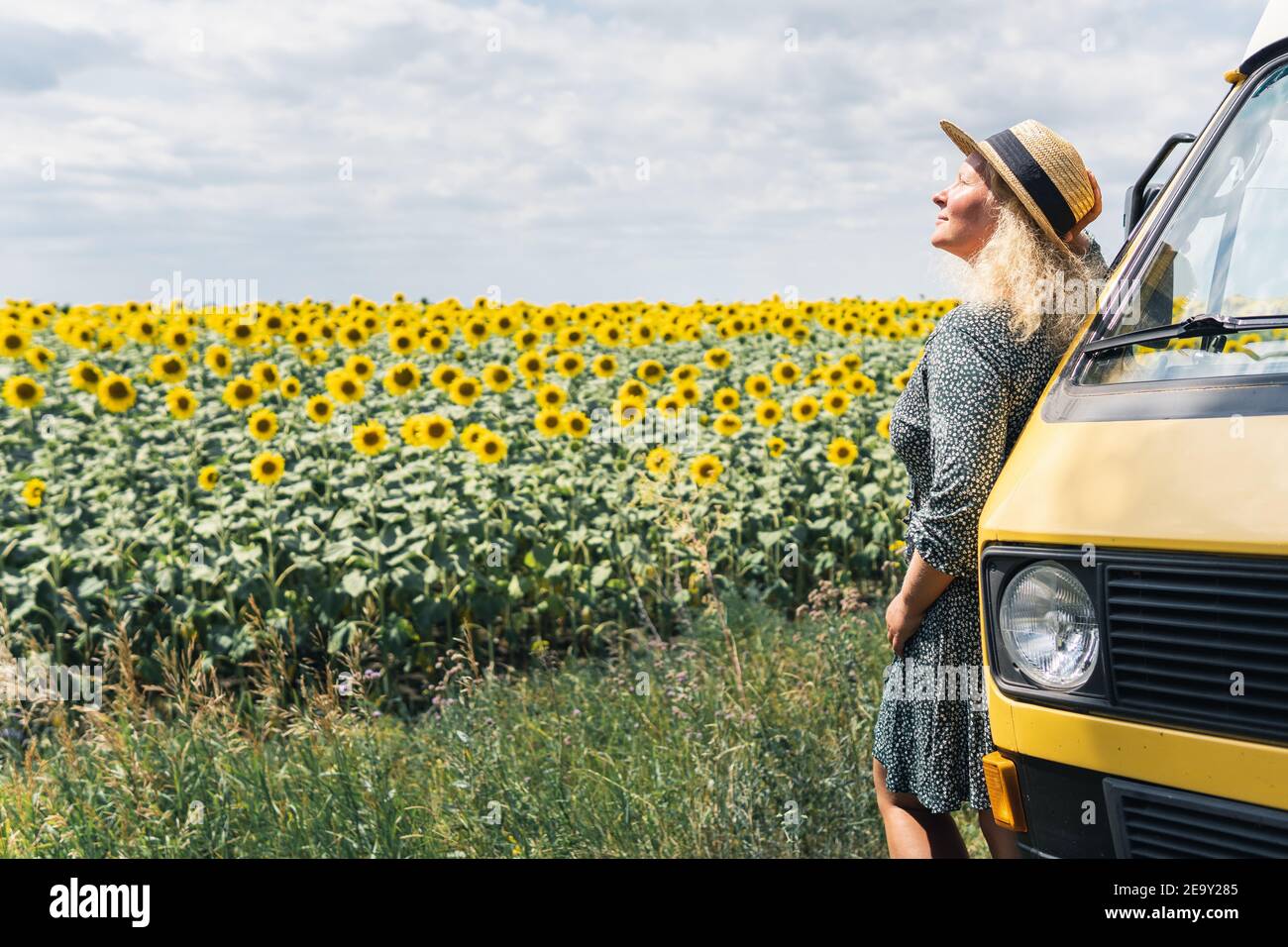 Junge blonde Frau, die mit dem Wohnmobil reist. Blick auf Sonnenblumenfeld an sonnigen Sommertag. Stockfoto