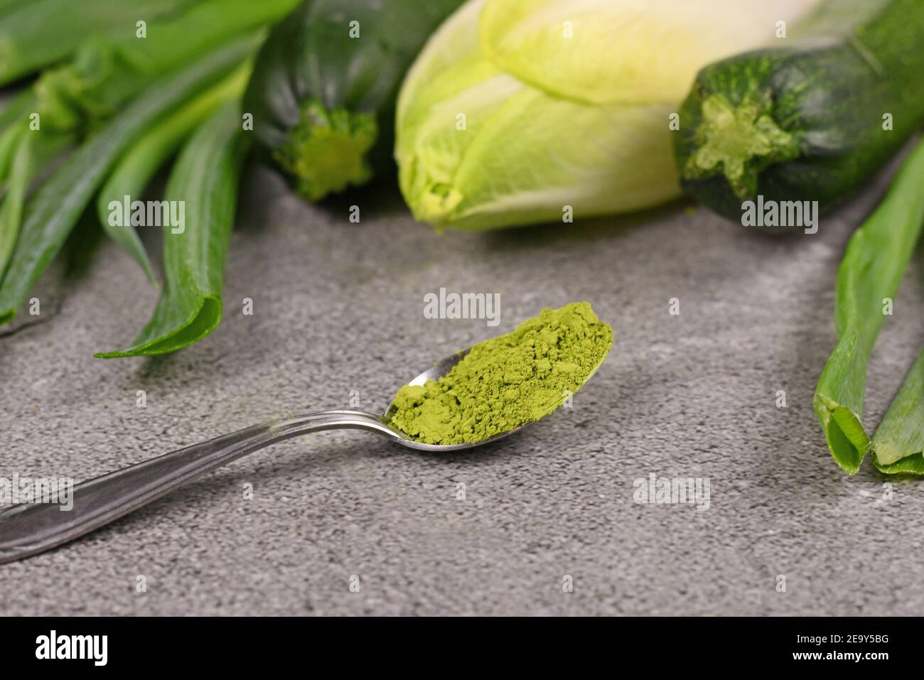 Grünes Pulver auf Löffel vor rohem grünem Gemüse. Konzept für natürliche Lebensmittelfarben oder Nahrungsergänzungsmittel aus Gemüse. Stockfoto