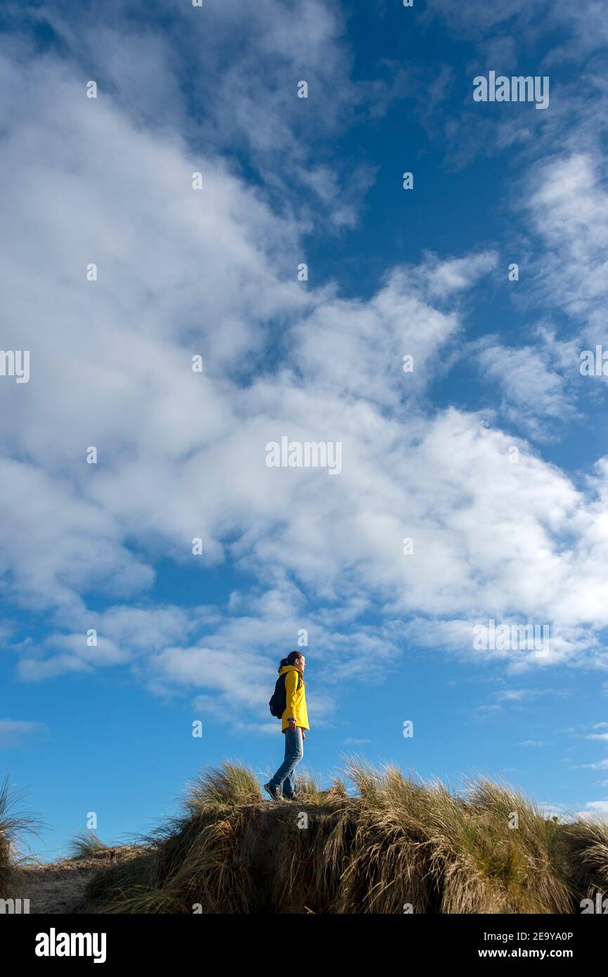 Weibliche Wanderin, die entlang hoher Sanddünen geht. Gelbe Jacke und Rucksack gegen einen blauen Himmel. Stockfoto