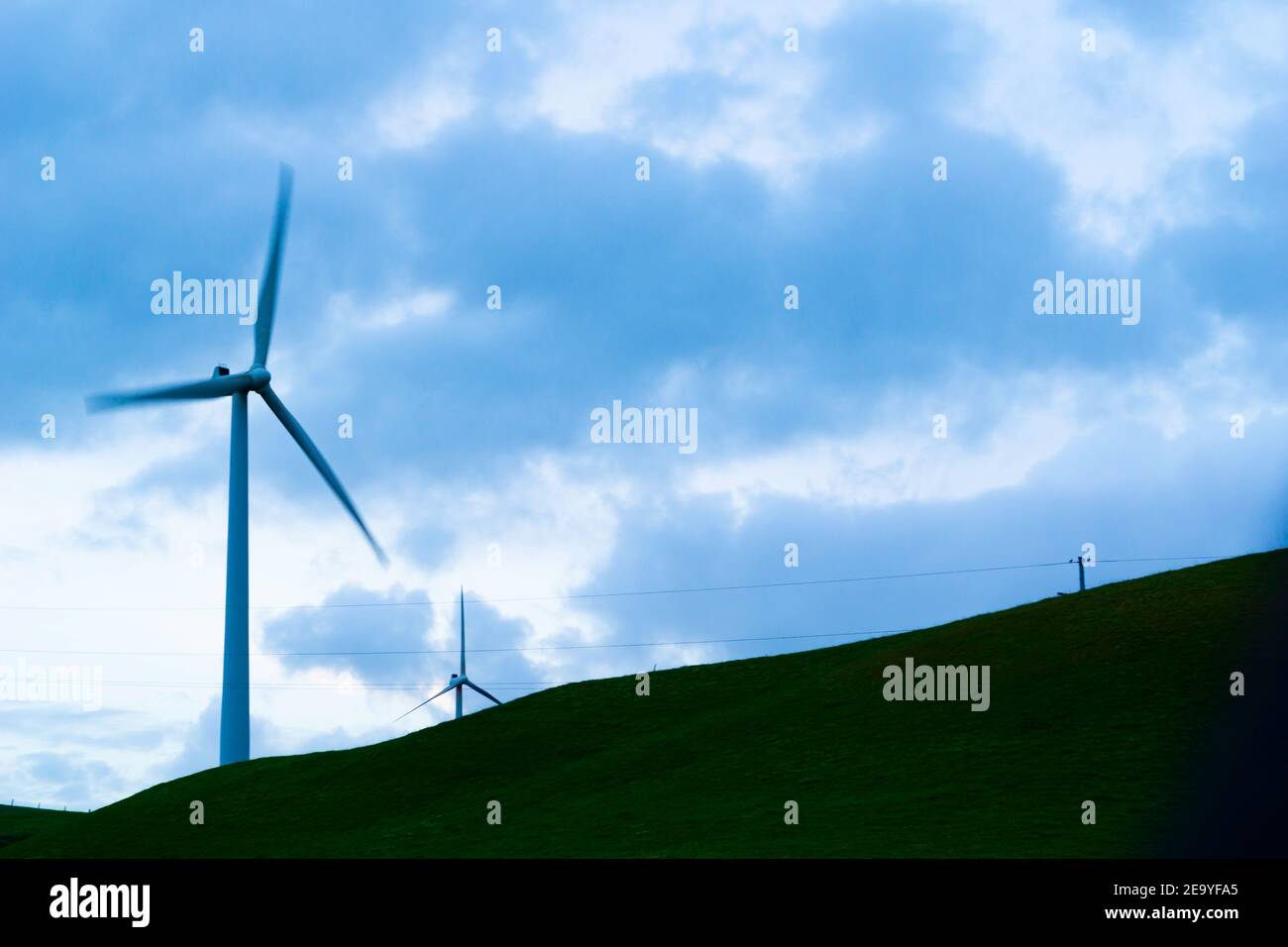 Verschwommene Rotorblätter oder Windturbinen, die sich langsam drehen, werden von untergehenden Sonnenstrahlen und bewölktem Himmel beleuchtet. Stockfoto