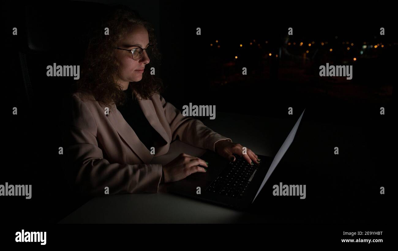 Frau im Anzug, die nachts arbeitet. Konzept der Überlastung im Büro, es ist dunkel und es gibt außerhalb des Fokus Lichter dahinter. Stockfoto