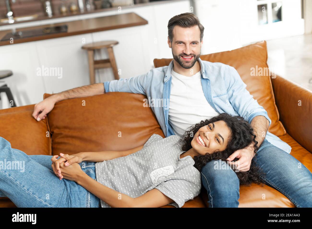 Lächelndes multiethnisches Paar in der Liebe ruht zu Hause zusammen, verbringt ein faules Wochenende auf der Couch im gemütlichen Wohnzimmer. Afrikanische Frau liegt auf den Schoß von Stockfoto