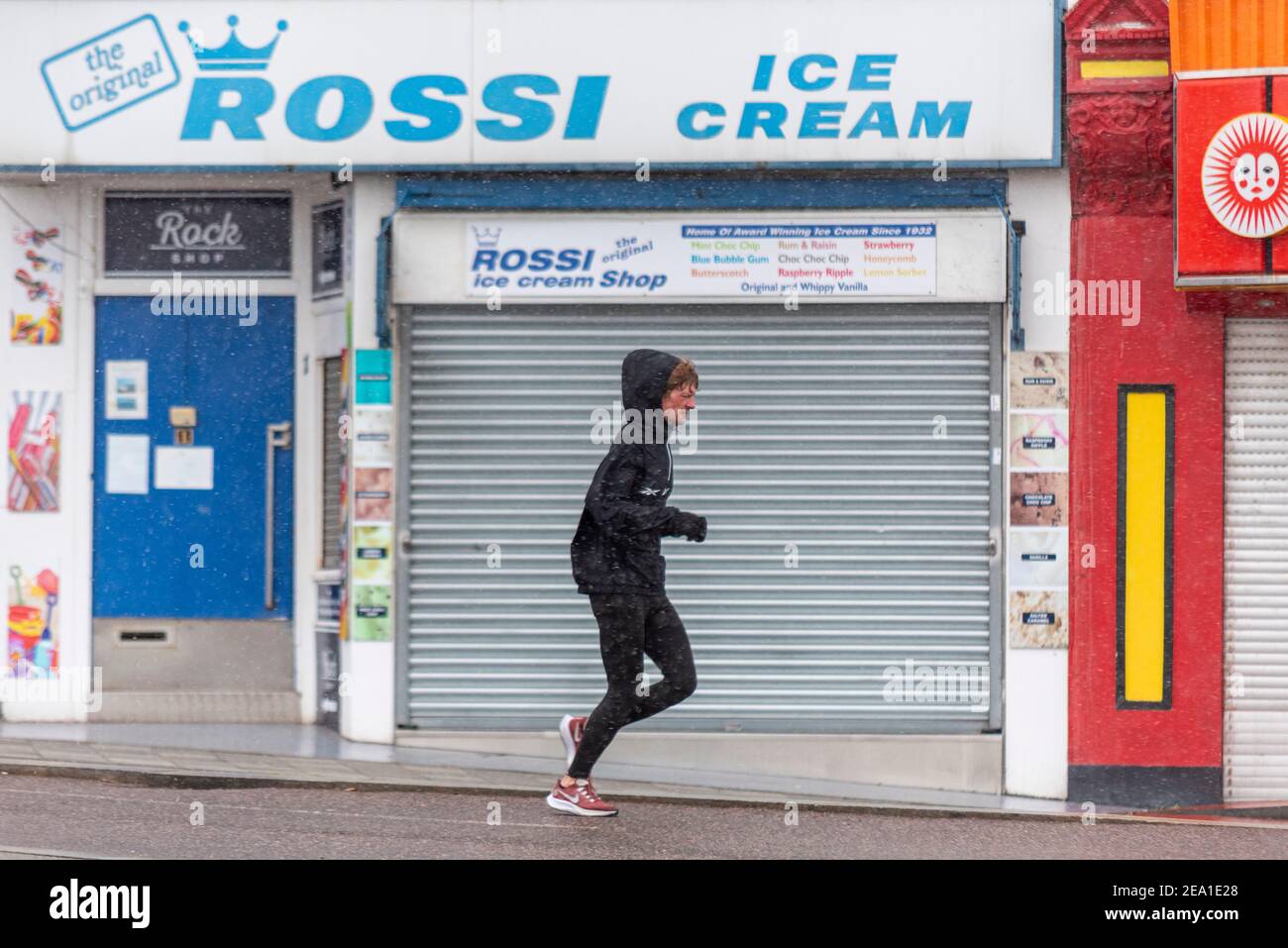 Southend on Sea, Essex, Großbritannien. Februar 2021, 7th. Storm Darcy erreichte Southend on Sea später als ursprünglich prognostiziert, wobei das Gebiet über Nacht nass blieb. Schnee und Schneestürmchen kamen um 7am an, was Jogger nicht abschreckte. COVID 19-Sperrwarnungen bleiben erhalten. Ein Rüde in Leggings und Kapuzenpulli läuft an geschlossenen Unternehmen am Meer vorbei. Geschlossen Rossi Eisdiele Stockfoto