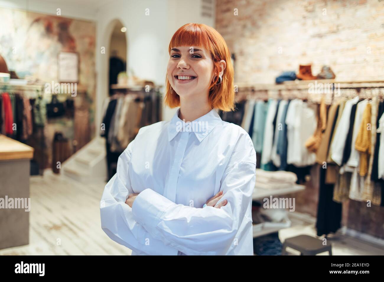 Porträt einer glücklichen jungen Frau, die mit gekreuzten Armen in ihrem Modehandel steht. Trendige weibliche Boutique-Besitzerin mit Blick auf die Kamera und lächelnd Stockfoto