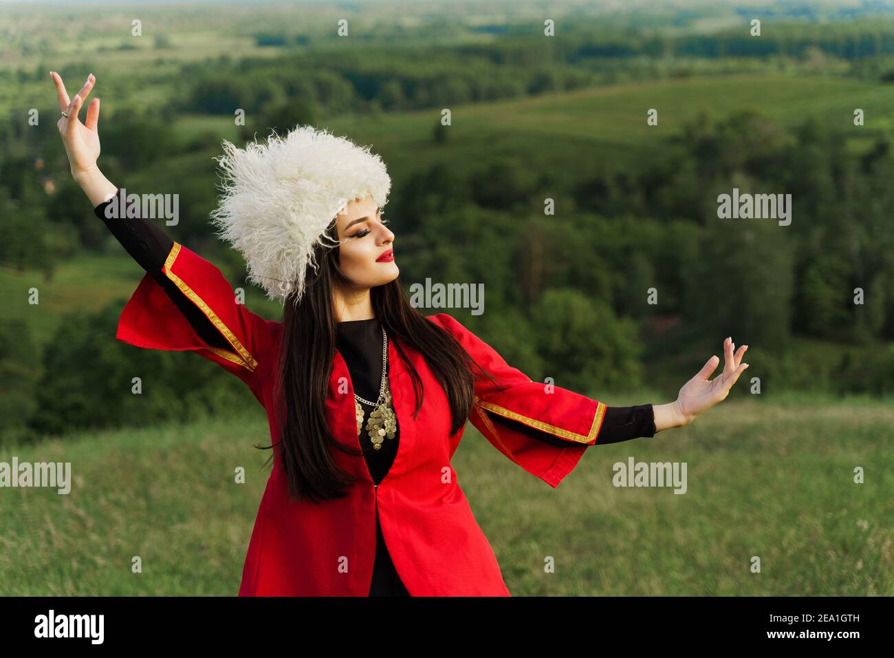 Georgische Mädchen in weißen Papakha Tänze Nationaltanz in roten Nationalkleid auf den grünen Hügeln von Georgien Hintergrund. Georgischer Kultur Lebensstil. Stockfoto