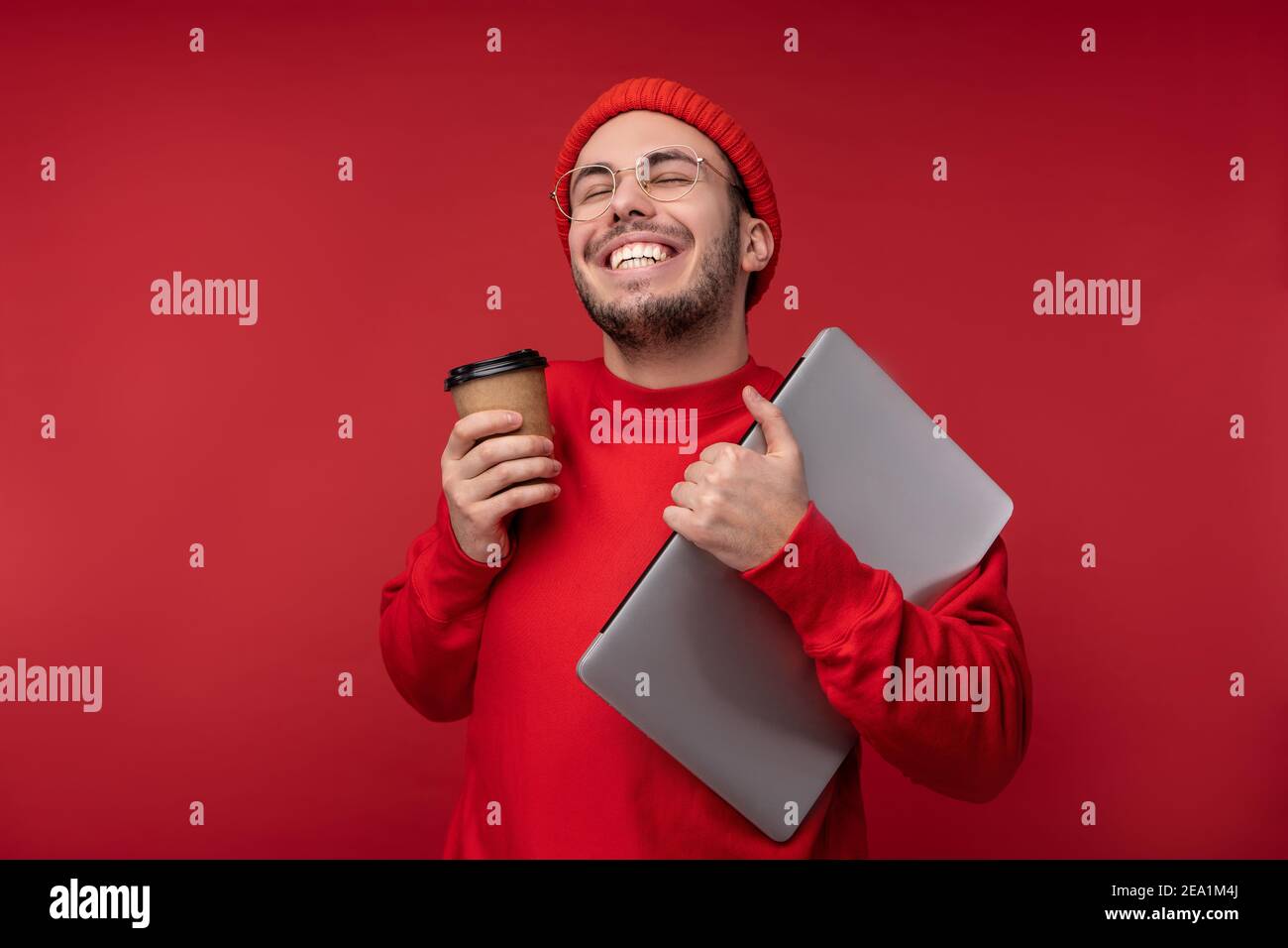 Foto von attraktiven Mann mit Bart in Brille und rote Kleidung. Glücklicher Mann hält Laptop und Kaffee fühlt sich gut an, isoliert auf rotem Hintergrund Stockfoto