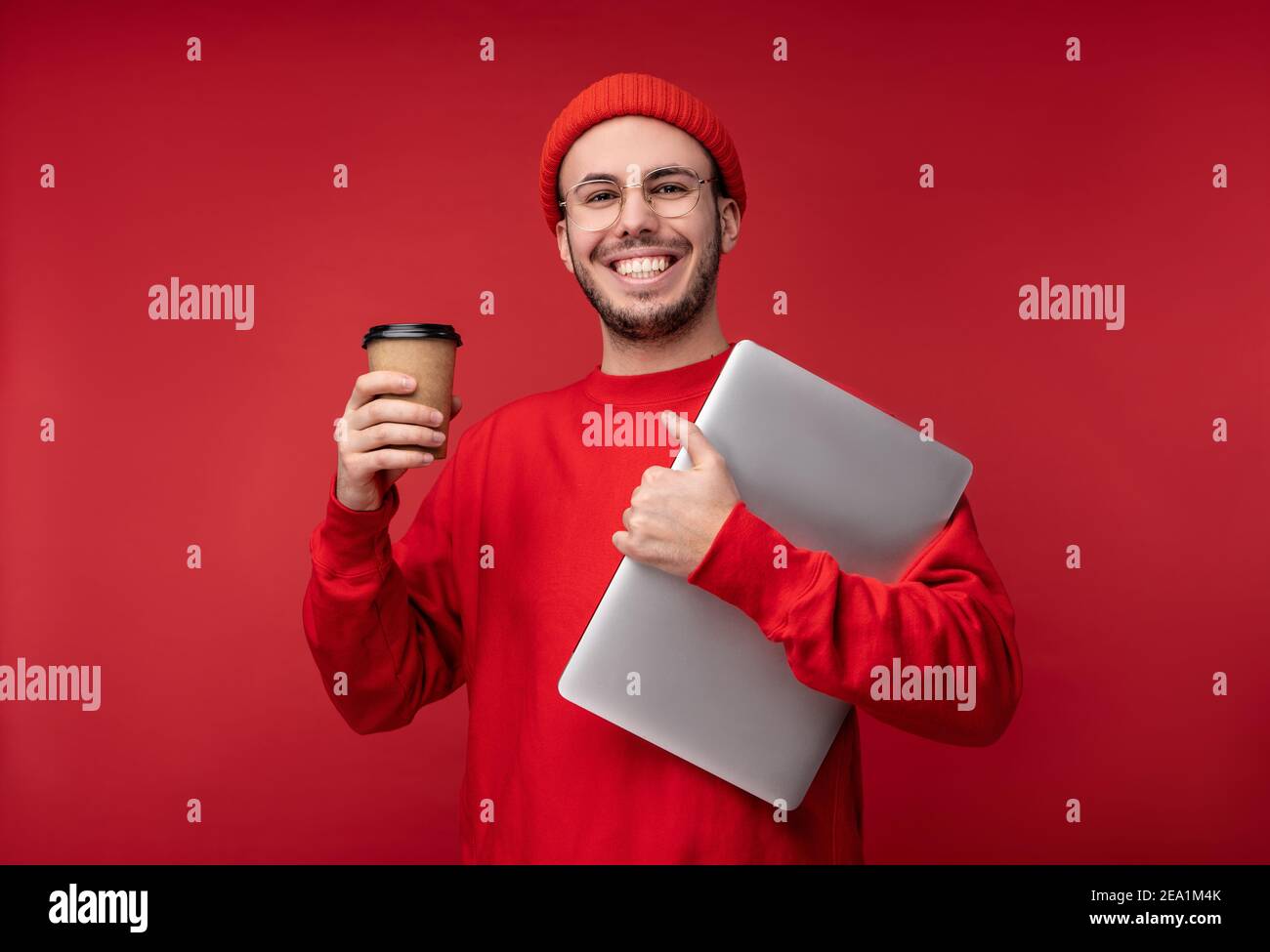 Foto von attraktiven Mann mit Bart in Brille und rote Kleidung. Glücklicher Mann hält Laptop und Kaffee fühlt sich gut an, isoliert auf rotem Hintergrund Stockfoto