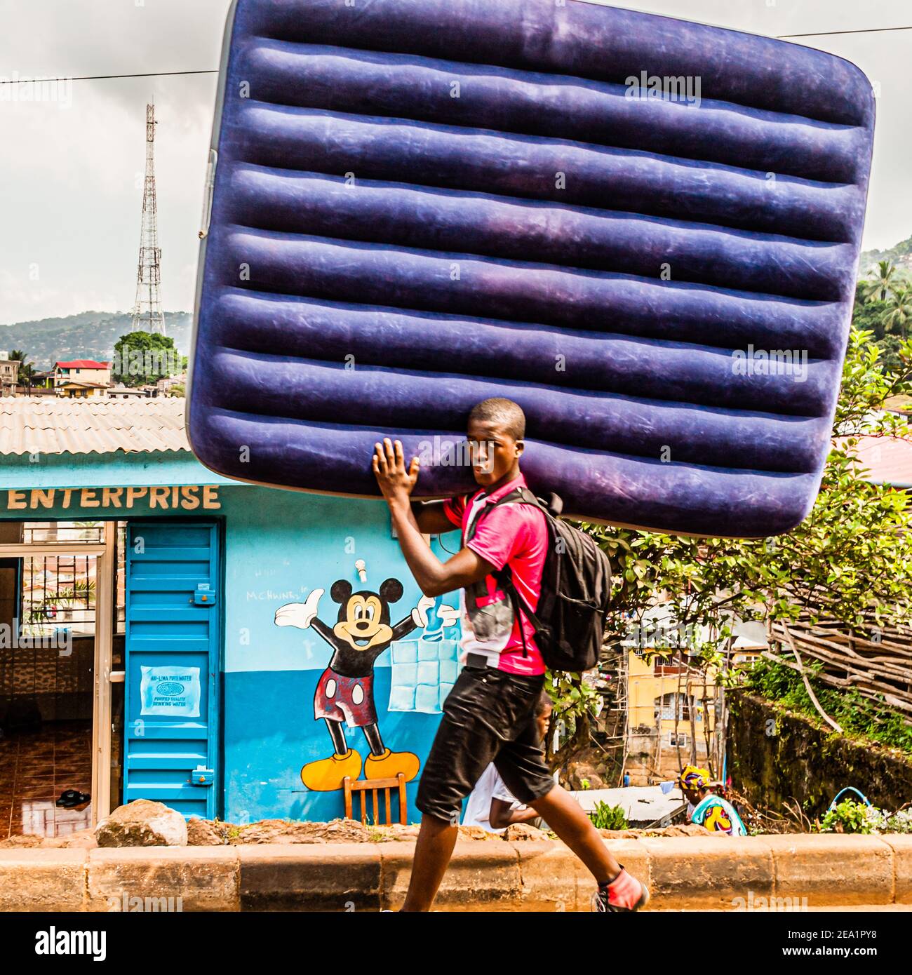 Der junge Mann transportiert eine aufgeblasene Luftmatratze vor einer Mickey-Maus-Firma in der westlichen Region Rural, Sierra Leone Stockfoto