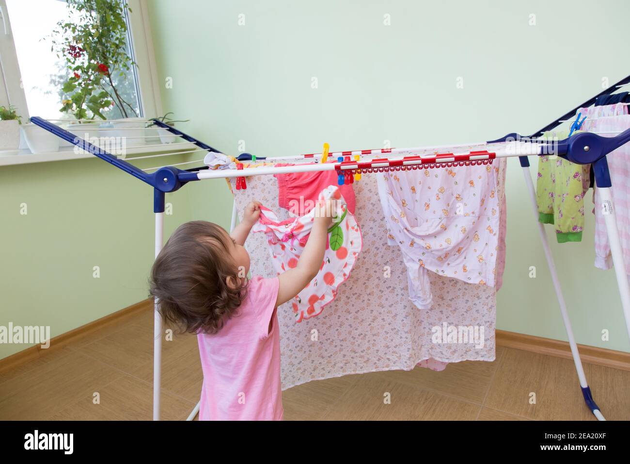 Babygirld hängt seine nassen Kleider zum Trocknen auf dem Balkon vor dem Hintergrund der Kalkwand. Konzept kleine Helfer Mama. Baby hält ihr helles d Stockfoto