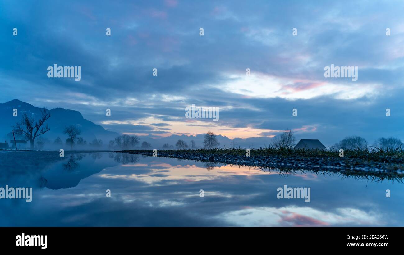 Sonnenuntergang mit Hütte, Baumspiegelung, Rheintal. Farbige Wolken und Schnee auf dem Feld. Farbige Wolken beim Sonnenuntergang im Rheintal, Baum Spiegelung Stockfoto