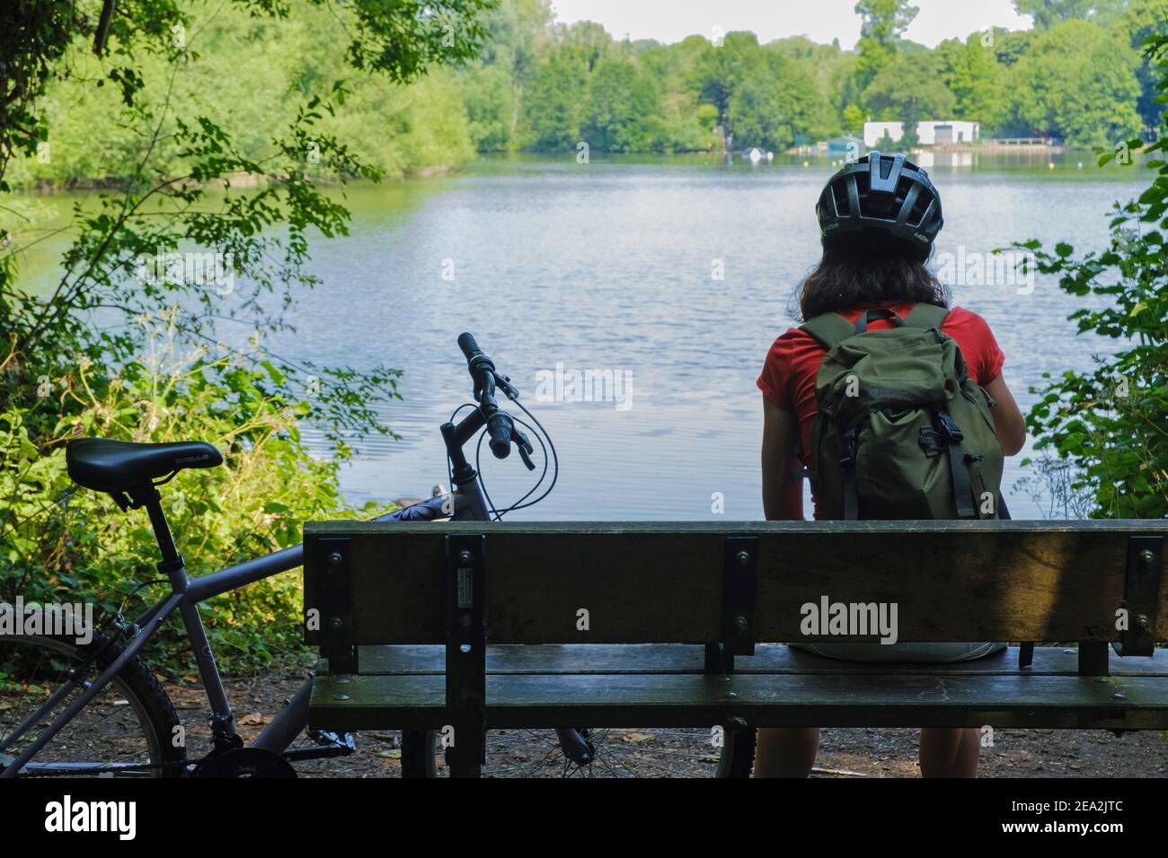 Rückansicht einer Dame mit Fahrradhelm & Rucksack auf einer Bank neben ihrem Fahrrad sitzend und dabei den Blick auf den Batchworth Lake, Rickmansworth Aquadrome geniesst. Stockfoto