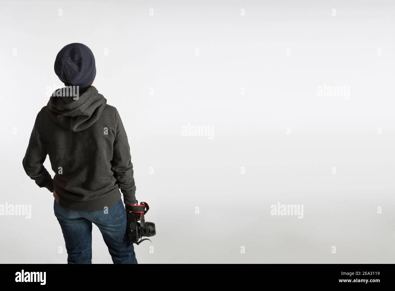 Ein Teenager mit einer Spiegelreflexkamera auf dem Rücken steht in einem Hoodie und einem Hut auf blauem Hintergrund. Schulungskonzept Fotografie Stockfoto