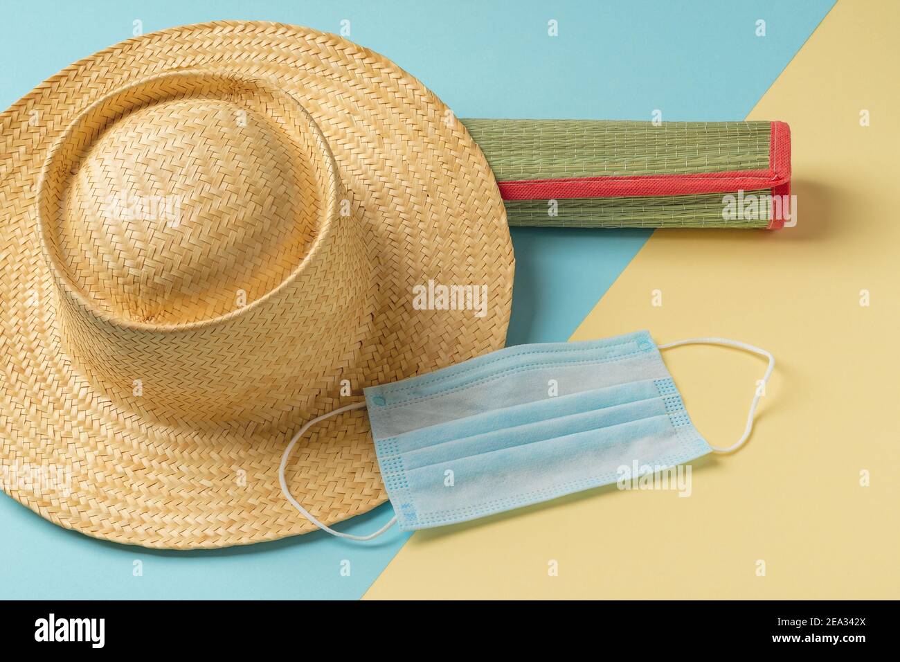 Medizinische Gesichtsmaske und Strohhut auf zusammengerollter Strandmatte vor gelbblauem Hintergrund. Schutz vor Kovid und Grippe während der Sommerferien, Reisen Stockfoto
