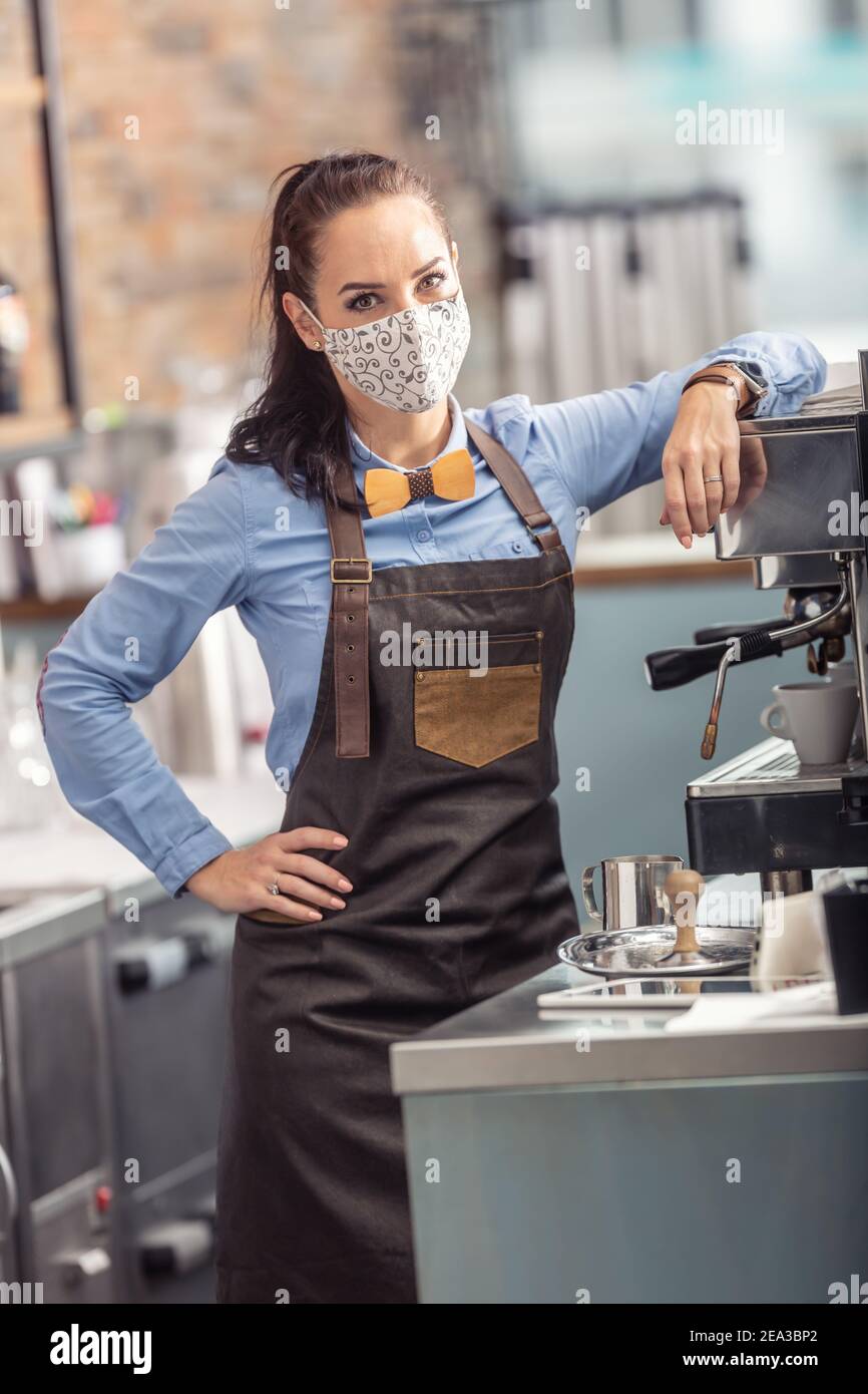 Die modische Kellnerin trägt eine schützende Gesichtsmaske, während sie einen Espresso in einem Kaffeehaus zubereitet. Stockfoto
